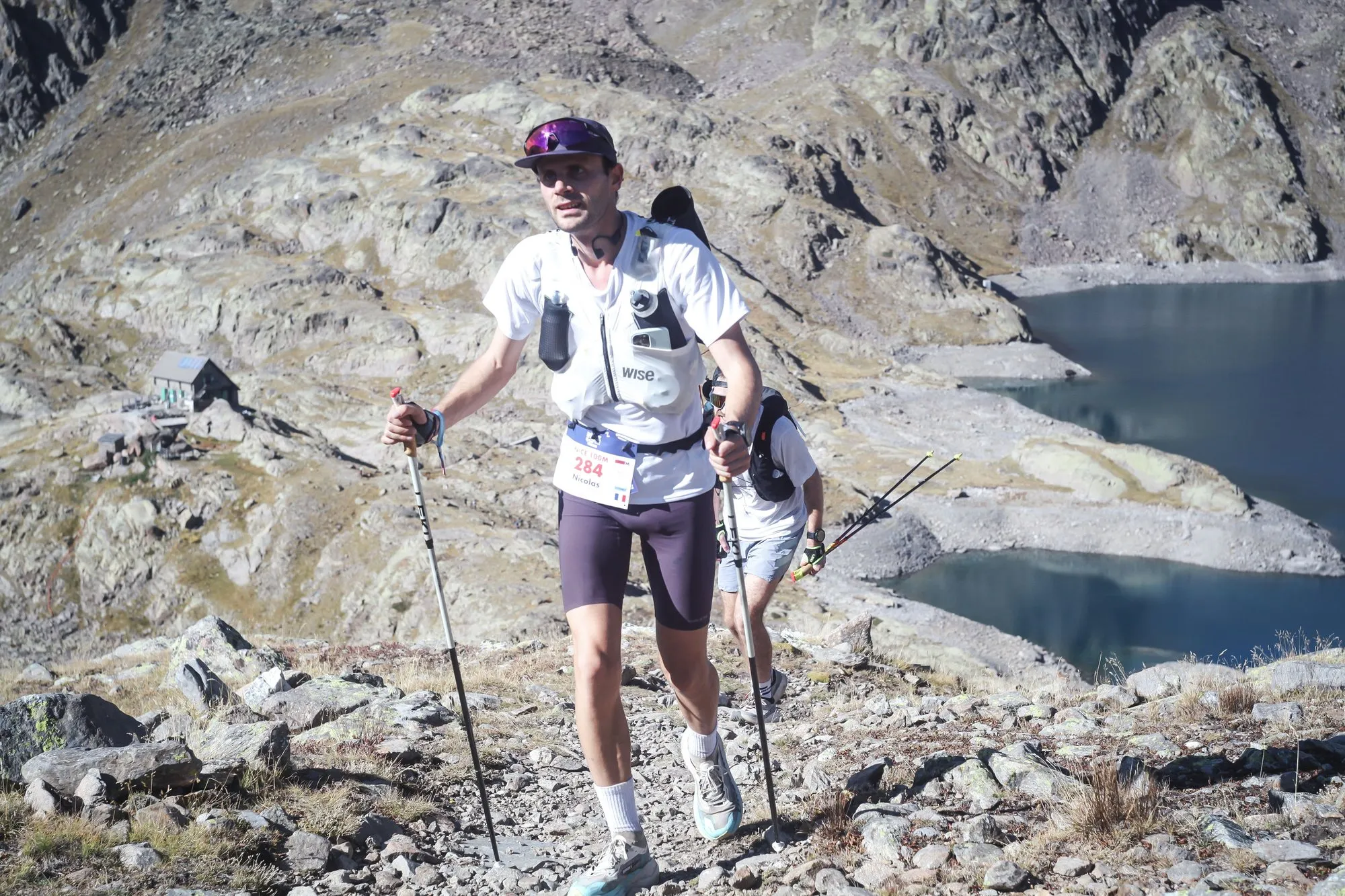 Nicolas Goullet de Rugy et un autre coureur sur un sentier caillouteux au-dessus d’un lac d’altitude, pendant l’UTMB Nice